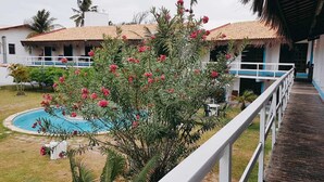 Vista de la piscina de un hotel con un edificio de techo de paja, una terraza de madera y un exuberante jardín con plantas en flor.