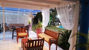 Un patio al aire libre con mobiliario de madera, incluyendo un banco y sillas, y una mesa. Hay una planta en maceta y una lámpara. El patio está cubierto por un dosel blanco y ofrece vistas a una piscina y follaje al fondo.