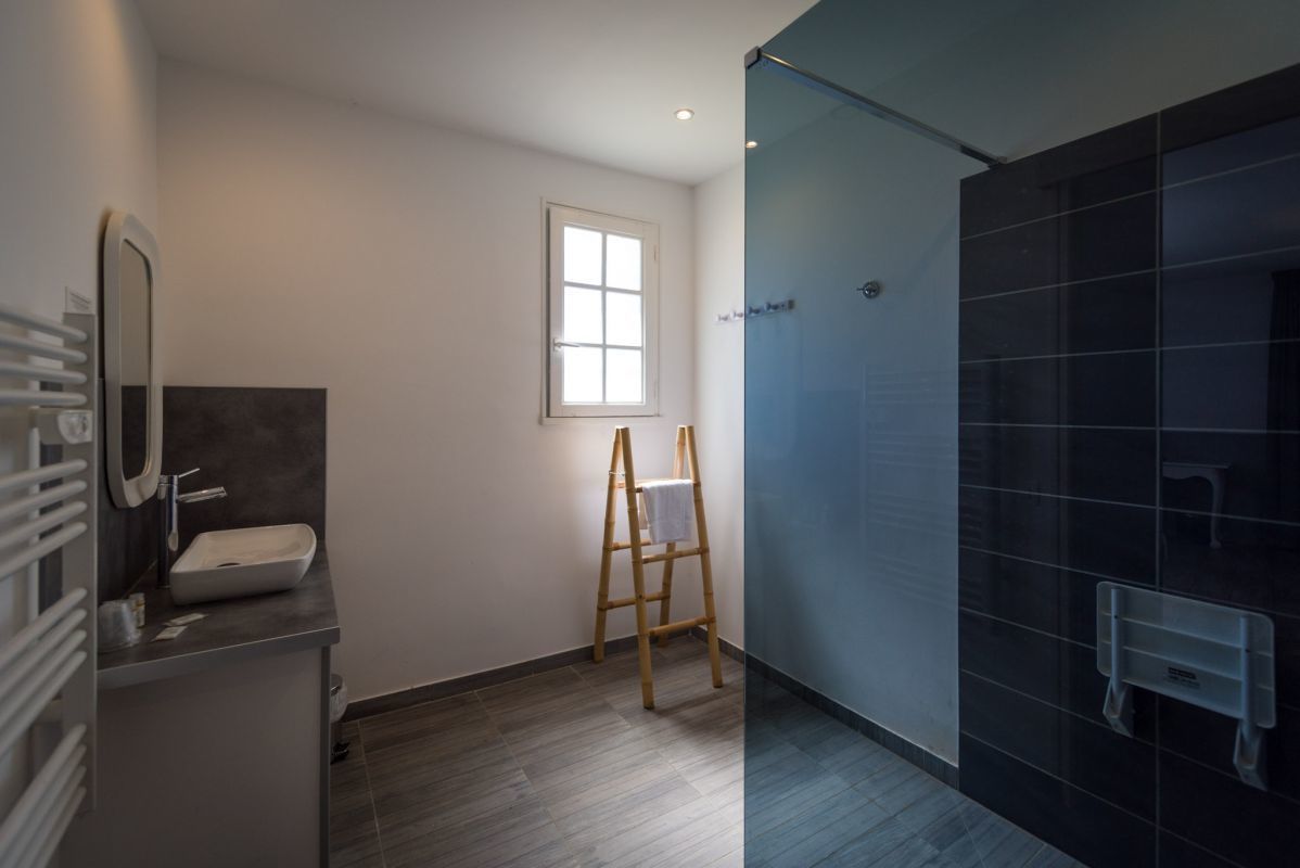 A modern bathroom with a dark tiled shower, a wooden ladder, a white sink, and a window providing natural light.