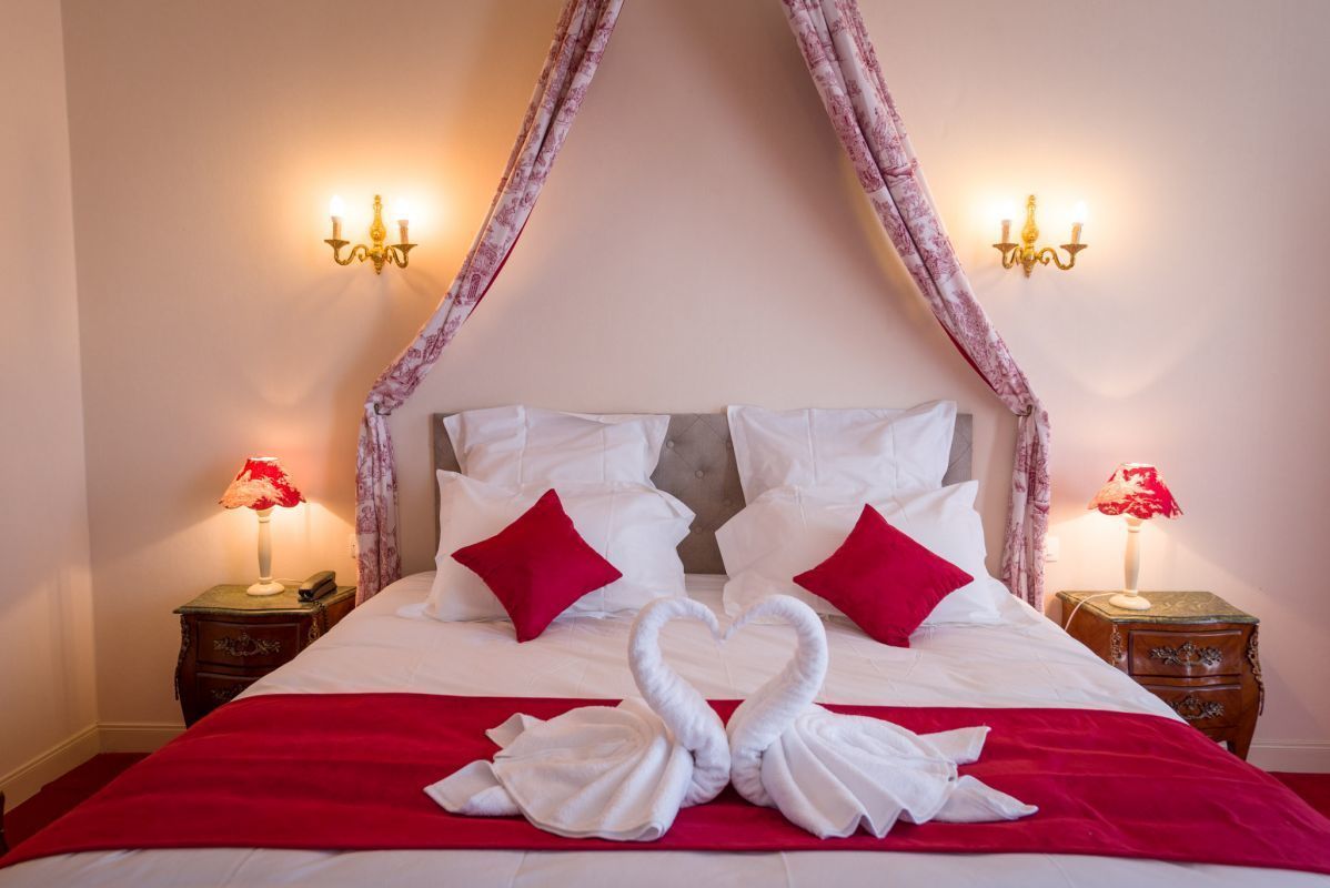 A bed with a canopy, white bedding, red pillows, and a red blanket. There are two bedside tables with lamps and a wall-mounted light fixture.