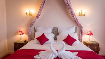 A bed with a canopy, white bedding, red pillows, and a red blanket. There are two bedside tables with lamps and a wall-mounted light fixture.