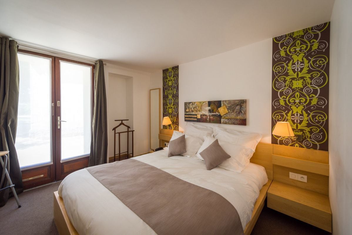 A hotel room with a large bed, a bedside table with a lamp, a window with curtains, and a wall with decorative patterns.