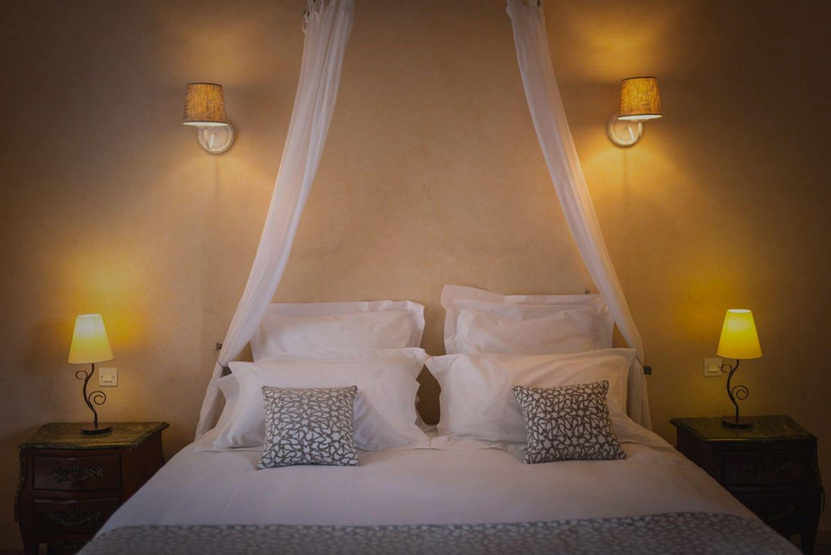 Una cama con ropa de cama blanca, dos almohadas estampadas y un dosel. Hay dos mesitas de noche con lámparas.