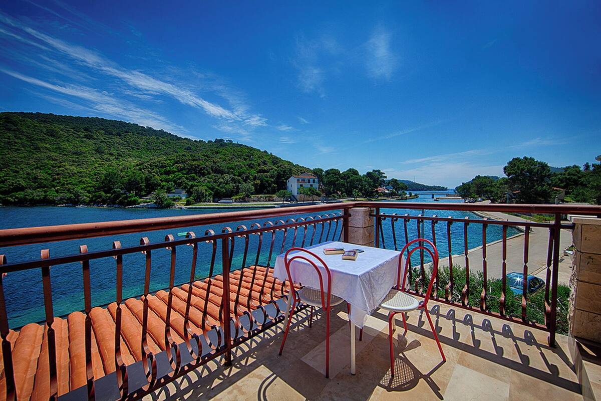 A balcony with a table and chairs overlooking a river, surrounded by lush greenery and a clear blue sky.