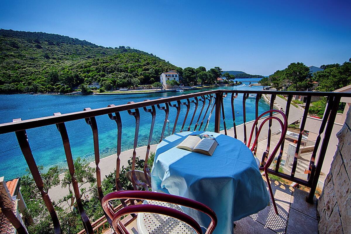 A balcony with a table and chairs overlooking a clear blue sea and a forested hillside.