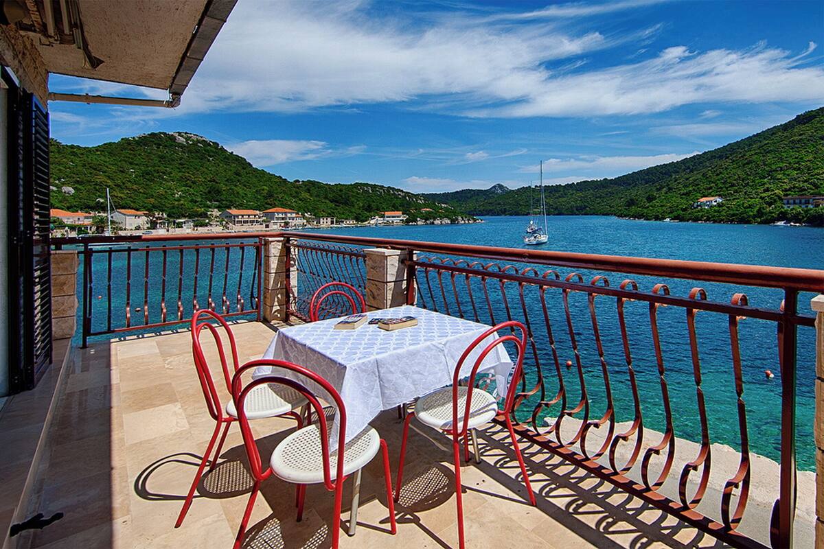 A balcony with a table and chairs, overlooking a body of water and a mountainous landscape.