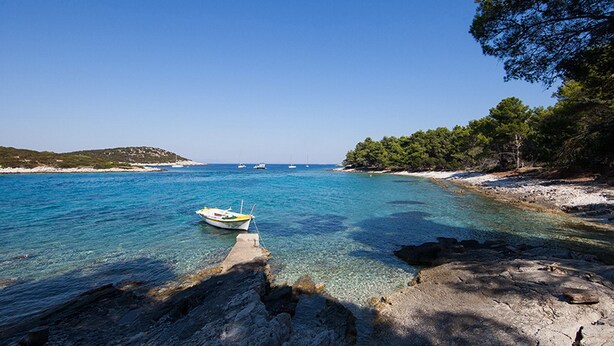 Un paysage côtier avec une eau turquoise et limpide, un petit bateau ancré près du rivage, un terrain rocheux et de nombreux arbres verdoyants.