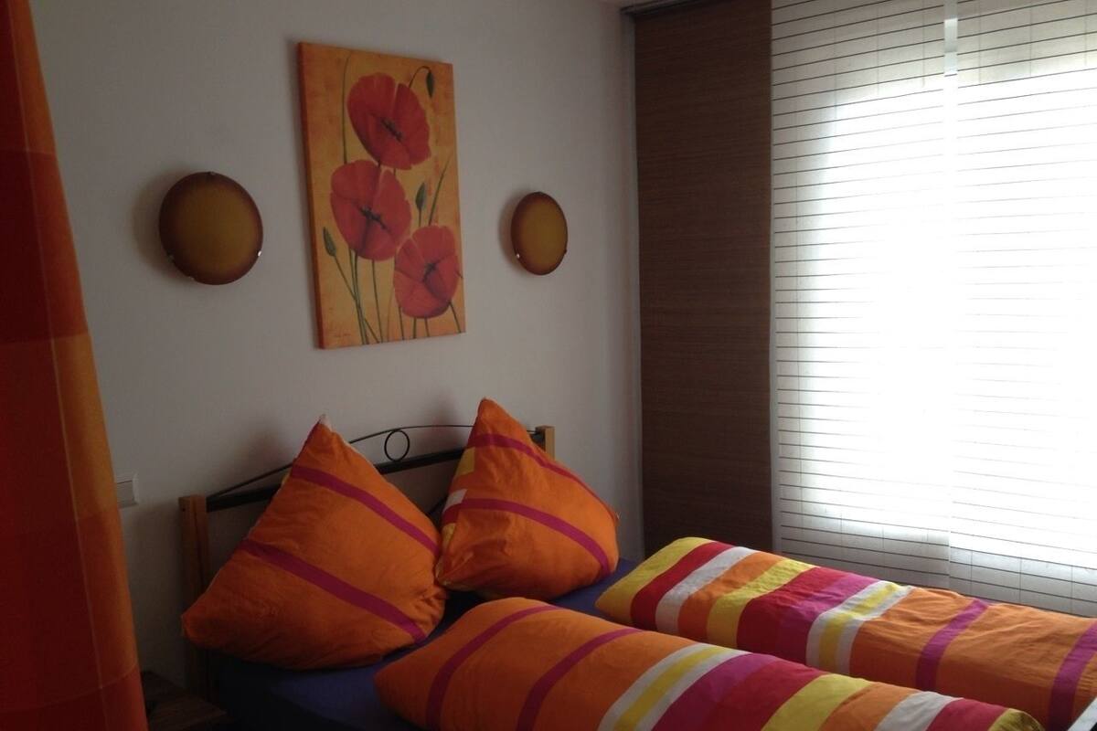 Une chambre avec deux lits, des oreillers orange, un tableau représentant des fleurs et une fenêtre avec des stores.