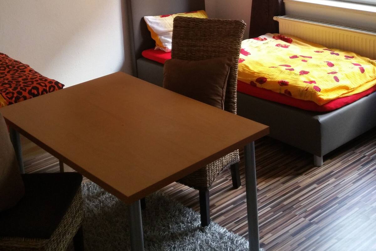Une chambre d’hôtel avec un lit, une table, une chaise, un tapis et une fenêtre avec des rideaux.