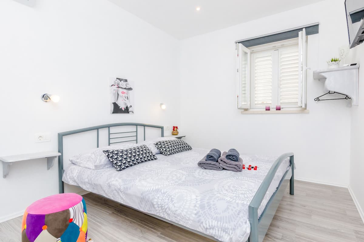 Une chambre bien rangée, avec un lit, des oreillers, un petit tabouret et une fenêtre avec des volets.