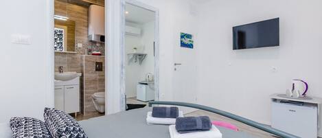 A modern hotel room with a bed, pillows, a TV, a desk, and a bathroom visible through an open door.