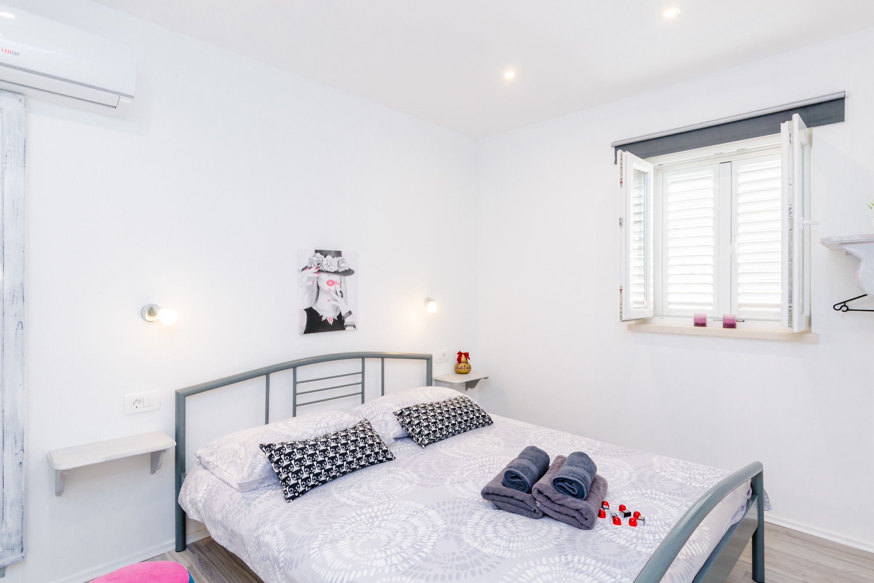 A bedroom with a bed, pillows, a nightstand, a window with shutters, and a wall-mounted shelf.