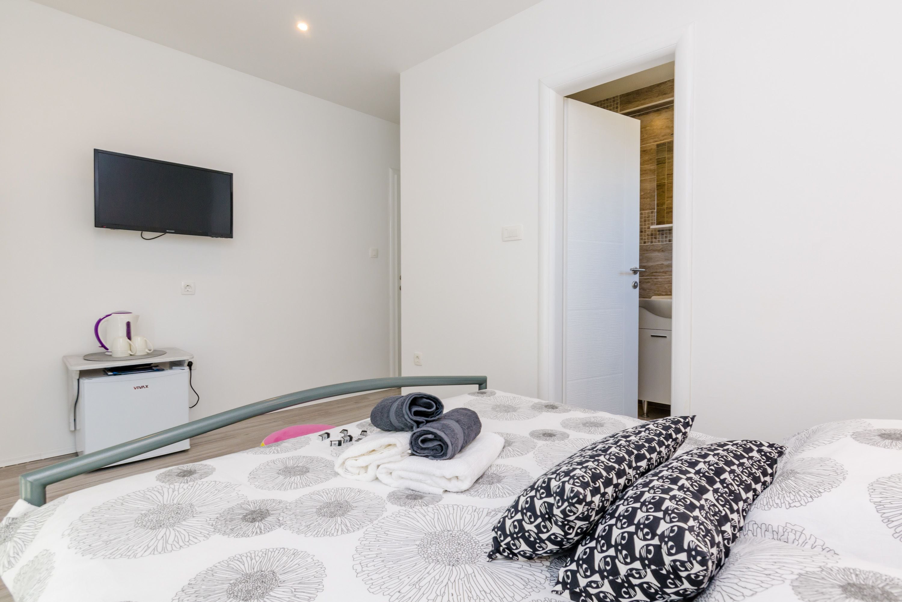 Ein modernes Schlafzimmer mit einem Bett, einem wandmontierten Flachbildfernseher, einem Nachttisch mit Wasserkocher und einer Tür zu einem Badezimmer.