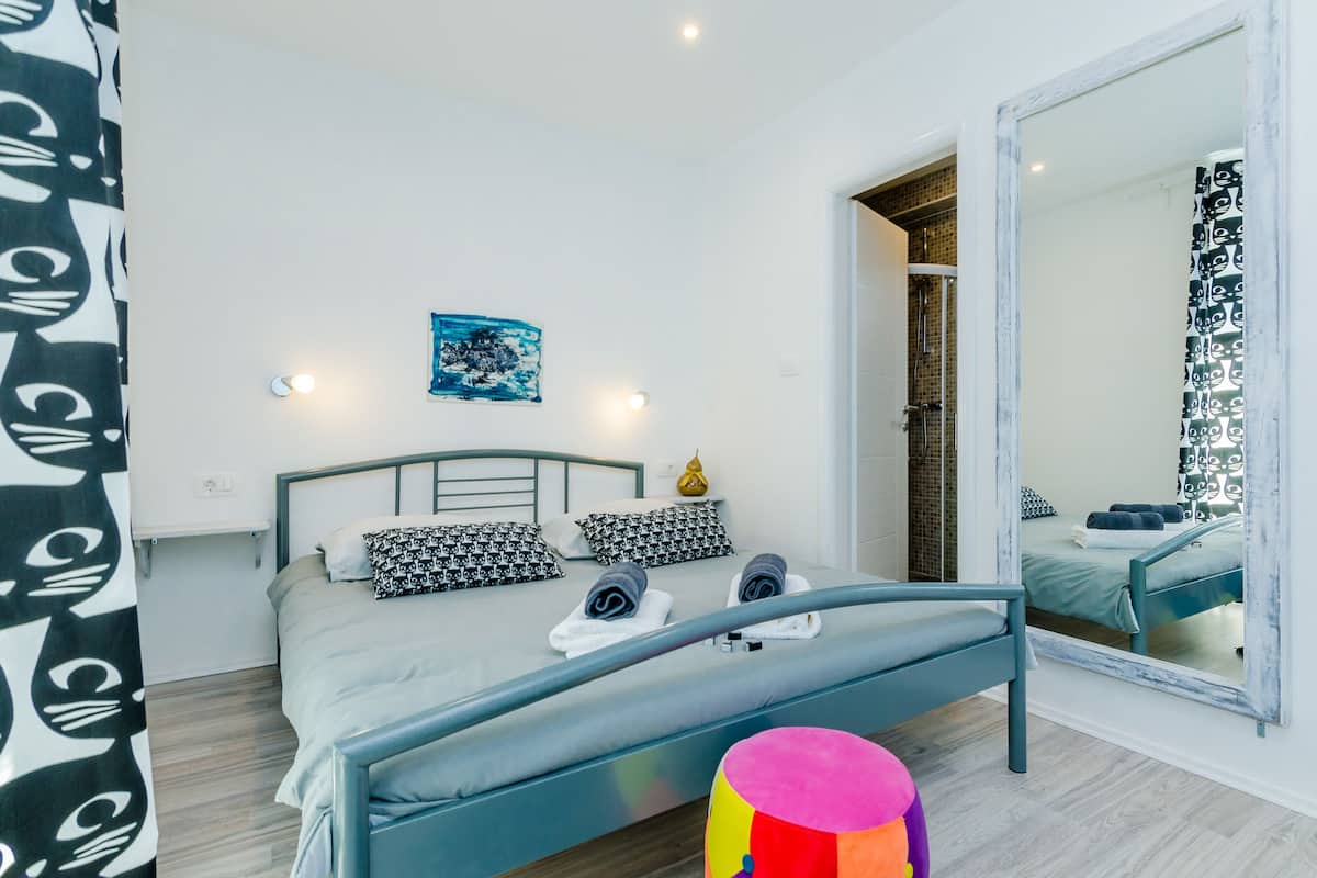 Une chambre à coucher moderne avec un lit, un tabouret coloré, un grand miroir et une petite œuvre d’art accrochée au mur.