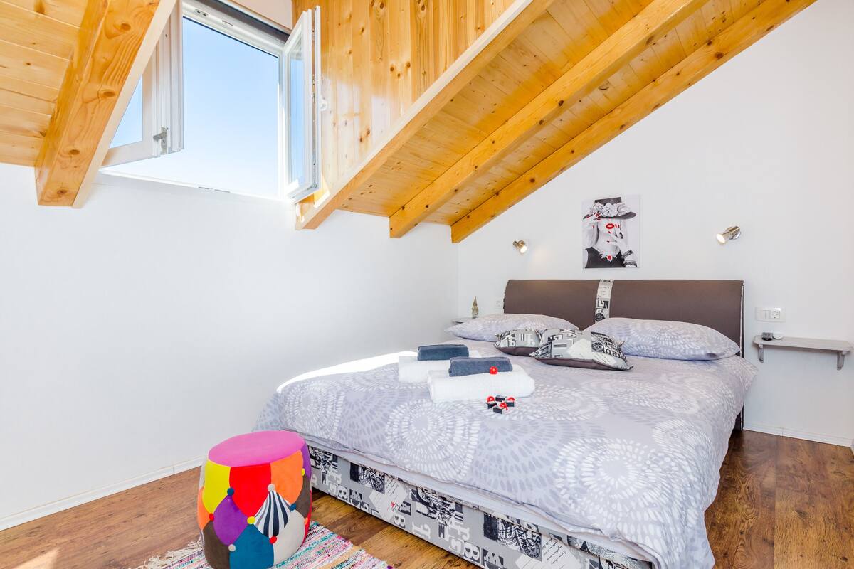 Une chambre avec un plafond en pente, un lit avec une literie à motifs, un tabouret coloré et une petite table sur laquelle est posé un objet décoratif.