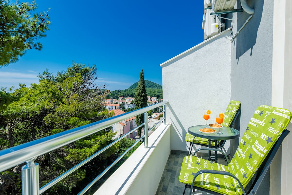Un balcon avec deux chaises vertes et une petite table, d’où l’on peut admirer un paysage urbain avec des immeubles et des arbres.