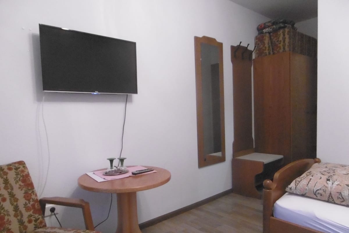 Une petite pièce comprenant un lit, une chaise, une table avec une télécommande, un téléviseur fixé au mur et un miroir.