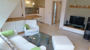 A modern living room with a sofa, a glass coffee table, a television, and a kitchen area in the background. - Antonia Rerik (Rerik)
