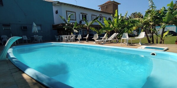 A swimming pool with lounge chairs and tables, surrounded by banana trees and a building with a tiled roof. - Pousada Canto dos Mares (Guarujá)