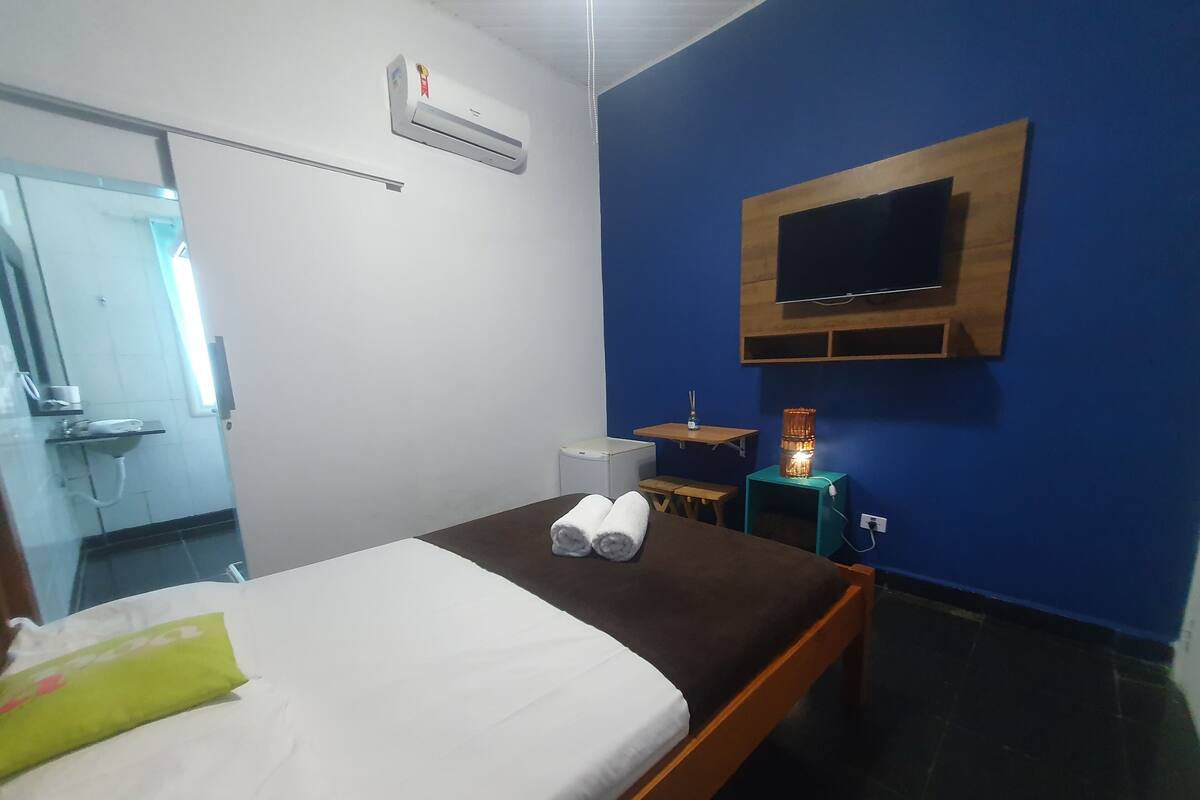 Habitación de hotel con una cama, un televisor montado en la pared, una mesita con una lámpara y un baño visible a través de una puerta abierta.