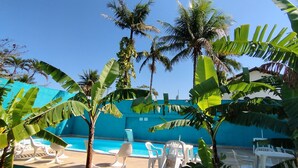 Zona junto a la piscina con sillas y mesas de plástico blanco, rodeada de palmeras y plantas de banana.
