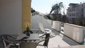 Une terrasse extérieure avec une table et des chaises, un pot de fleurs et un portail blanc donnant sur un escalier.