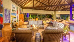 Un amplio espacio lounge al aire libre con mobiliario de mimbre, techo de paja y cuadros en las paredes.