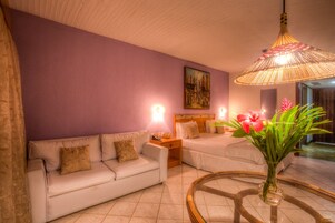 Habitación de hotel con pared morada, dos camas, un sofá blanco, una mesa de centro de vidrio y una lámpara colgante.