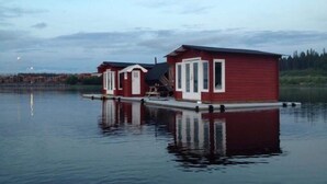 Zwei rote schwimmende Häuser mit weißem Verzierstreifen, verbunden durch eine Anlegestelle, auf einem ruhigen See.