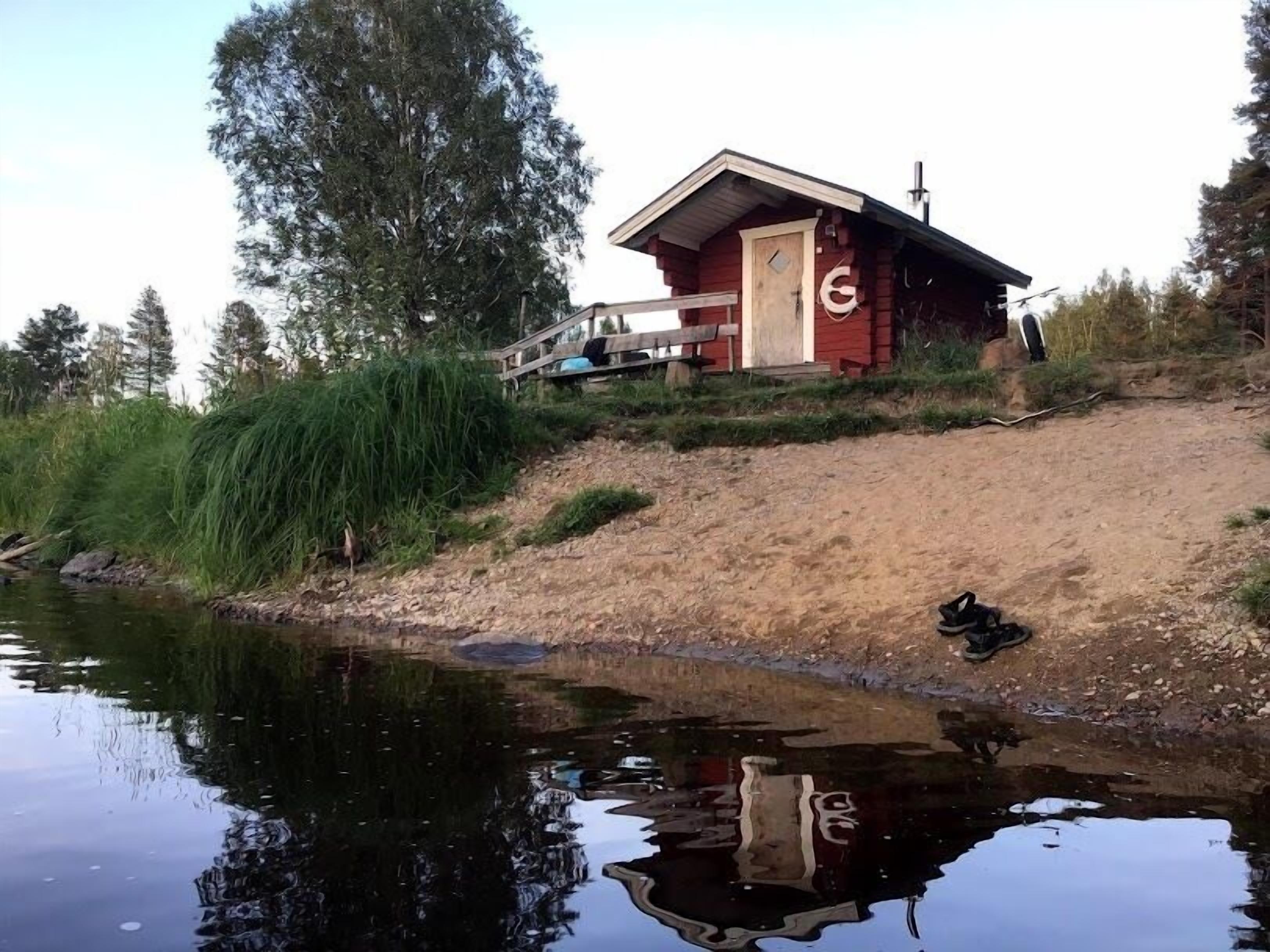 A small cabin with a wooden door and a 'G' sign, situated beside a calm body of water with reflections.