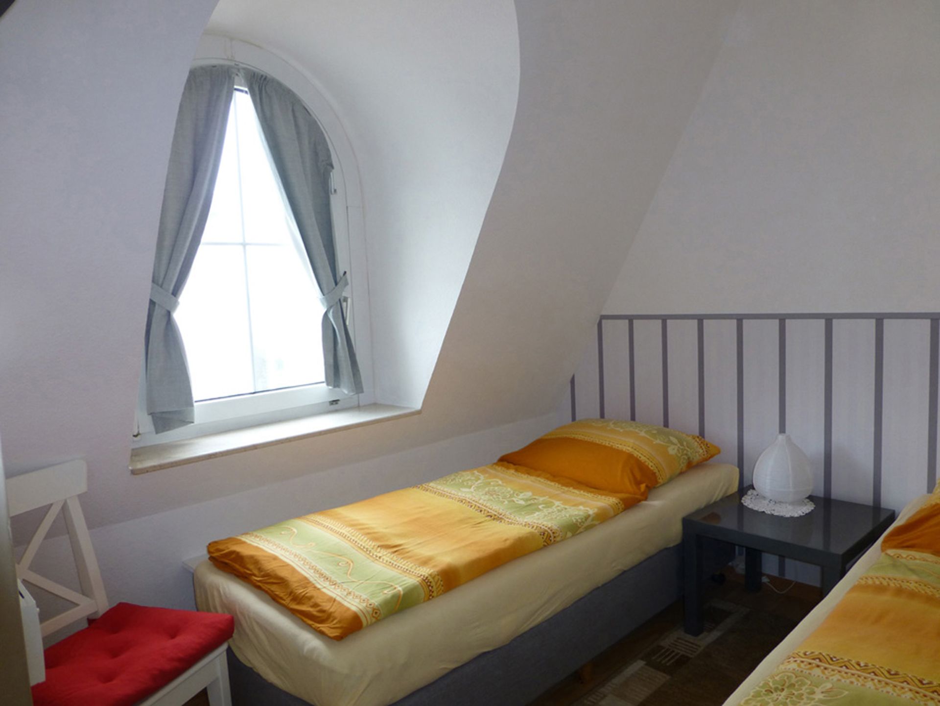 Une petite chambre avec un lit simple, une fenêtre, une table de chevet avec une lampe et une chaise rouge.