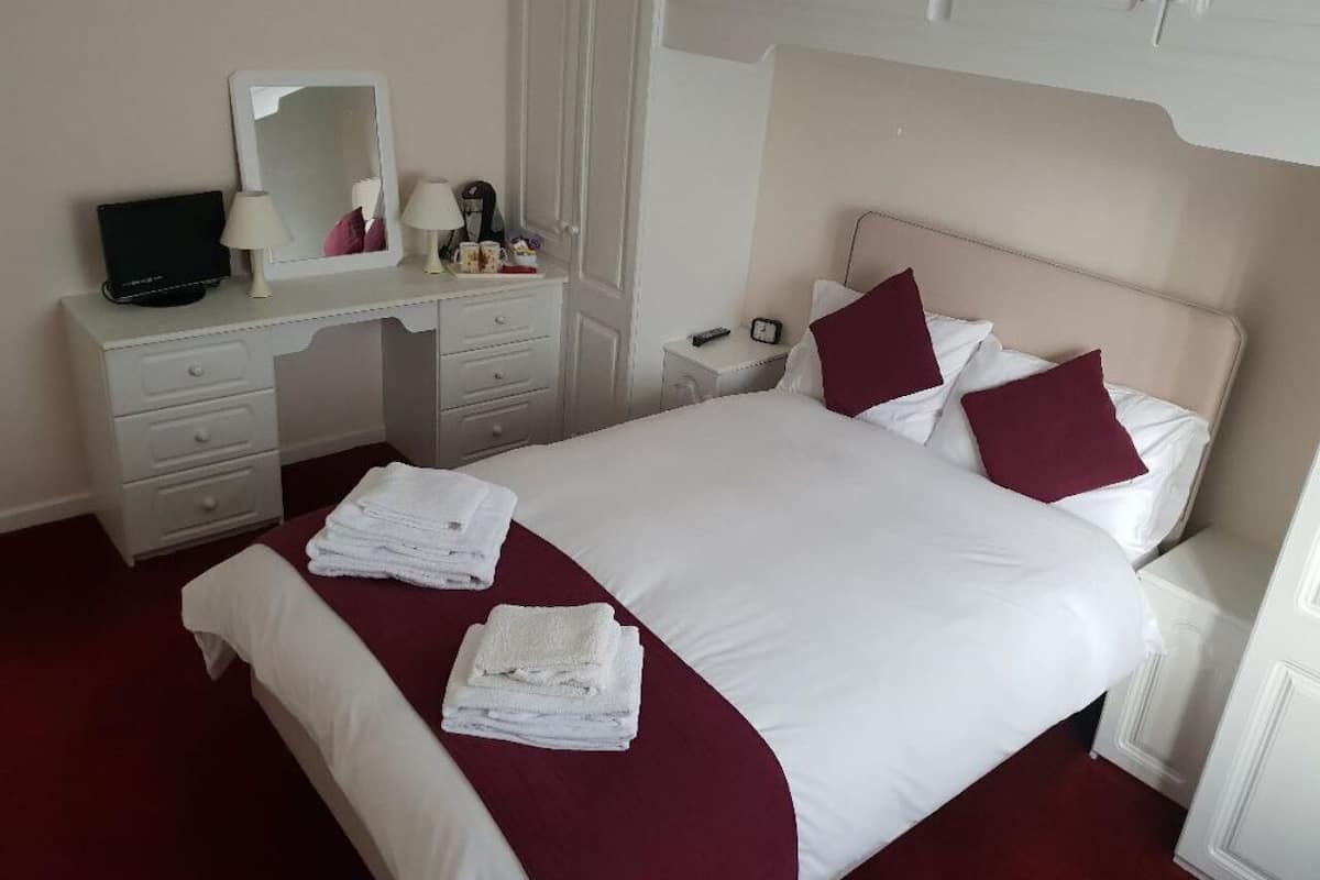 A neatly made bed with white and maroon bedding, a bedside table with a lamp, a mirror, and a small TV.