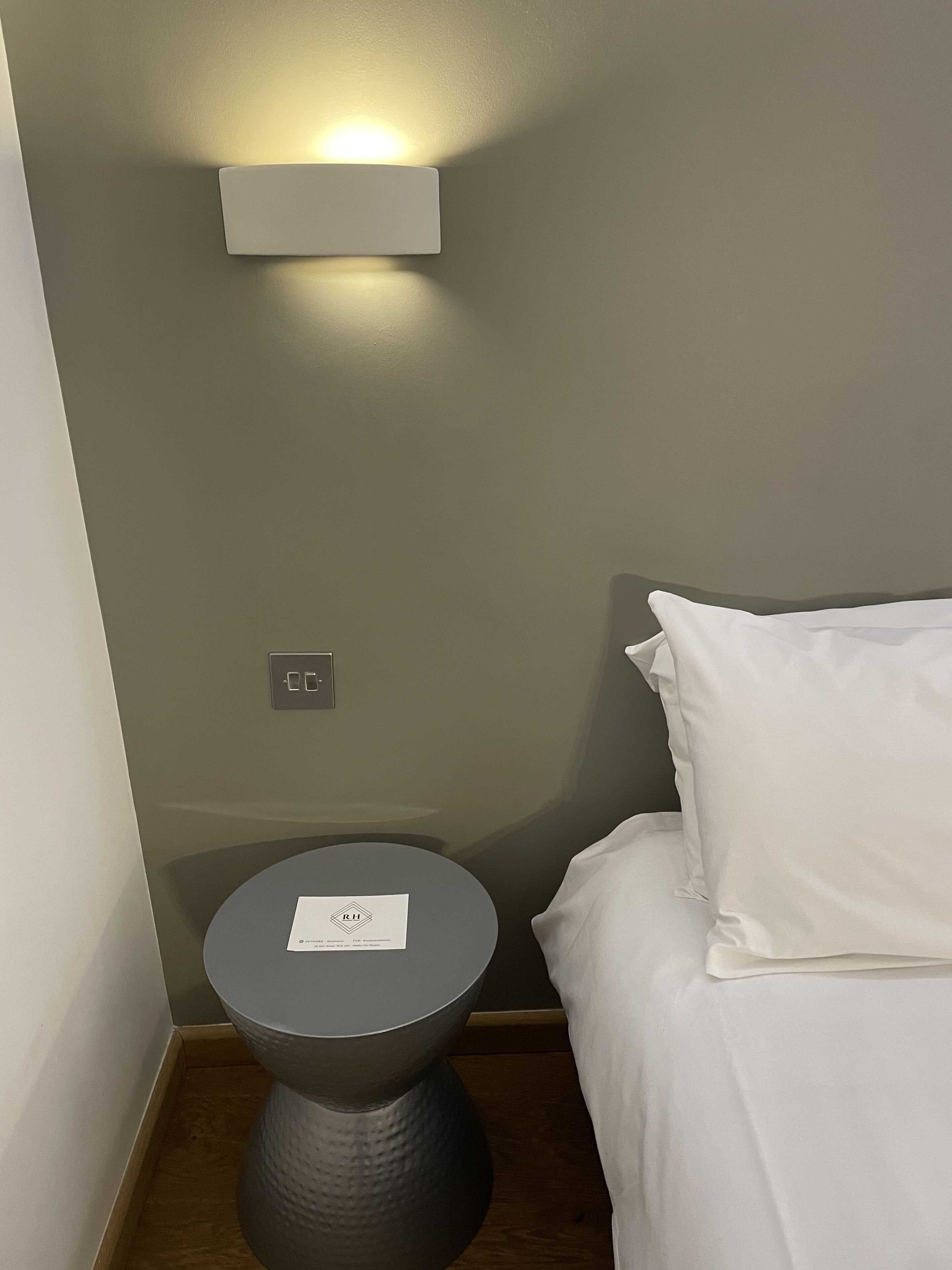 Una mesita auxiliar pequeña, redonda y oscura con una etiqueta, un almohadón blanco y una cama en una habitación con pared de color claro y una lámpara pequeña colgante sobre la mesa.