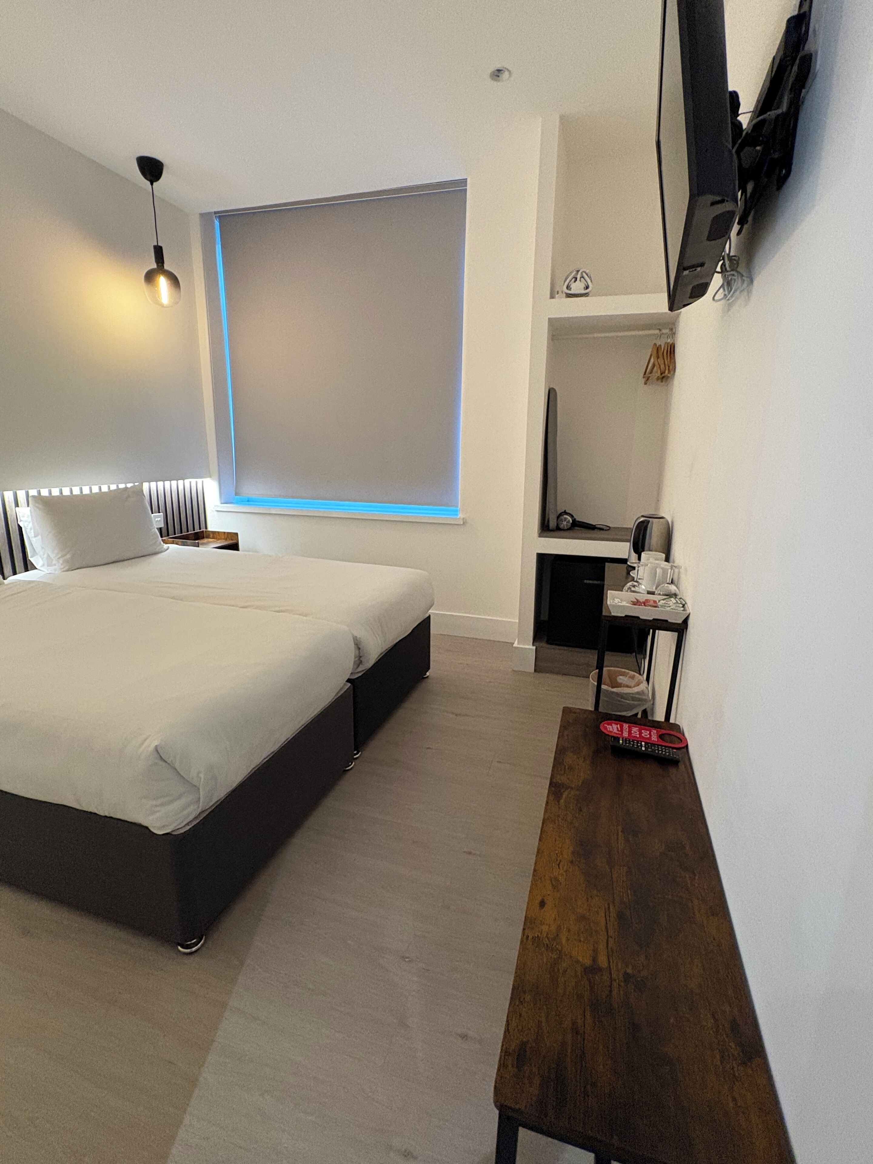 A modern hotel room with a large bed, a desk with a computer, a TV mounted on the wall, and a window with blinds.