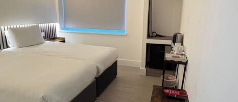 A modern hotel room with a large bed, a desk with a computer, a TV mounted on the wall, and a window with blinds.