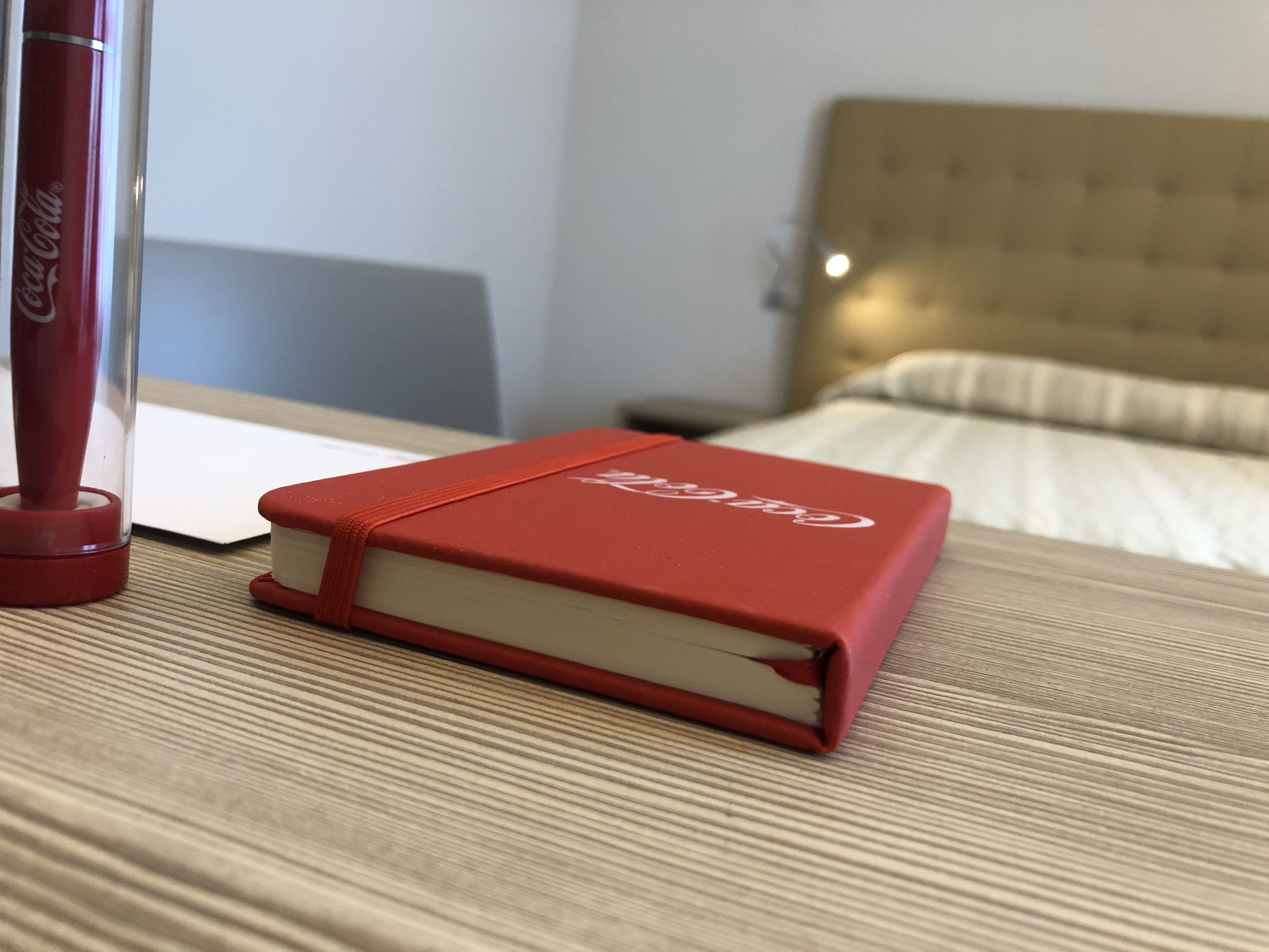 Una libreta roja con el logotipo de Coca-Cola sobre una superficie de madera, un bolígrafo rojo y una cama con cabecero color beige al fondo.