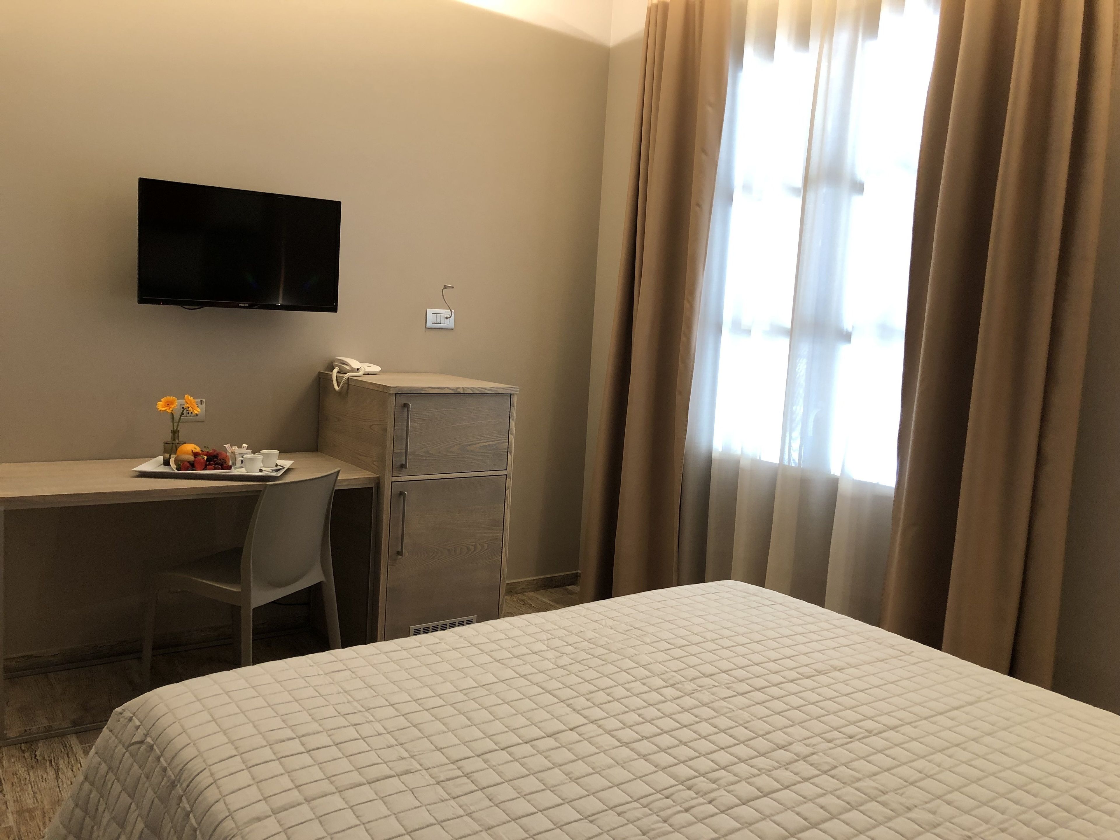 Una camera d'albergo con un letto, una scrivania con una sedia, una televisione a muro e una finestra con tende.