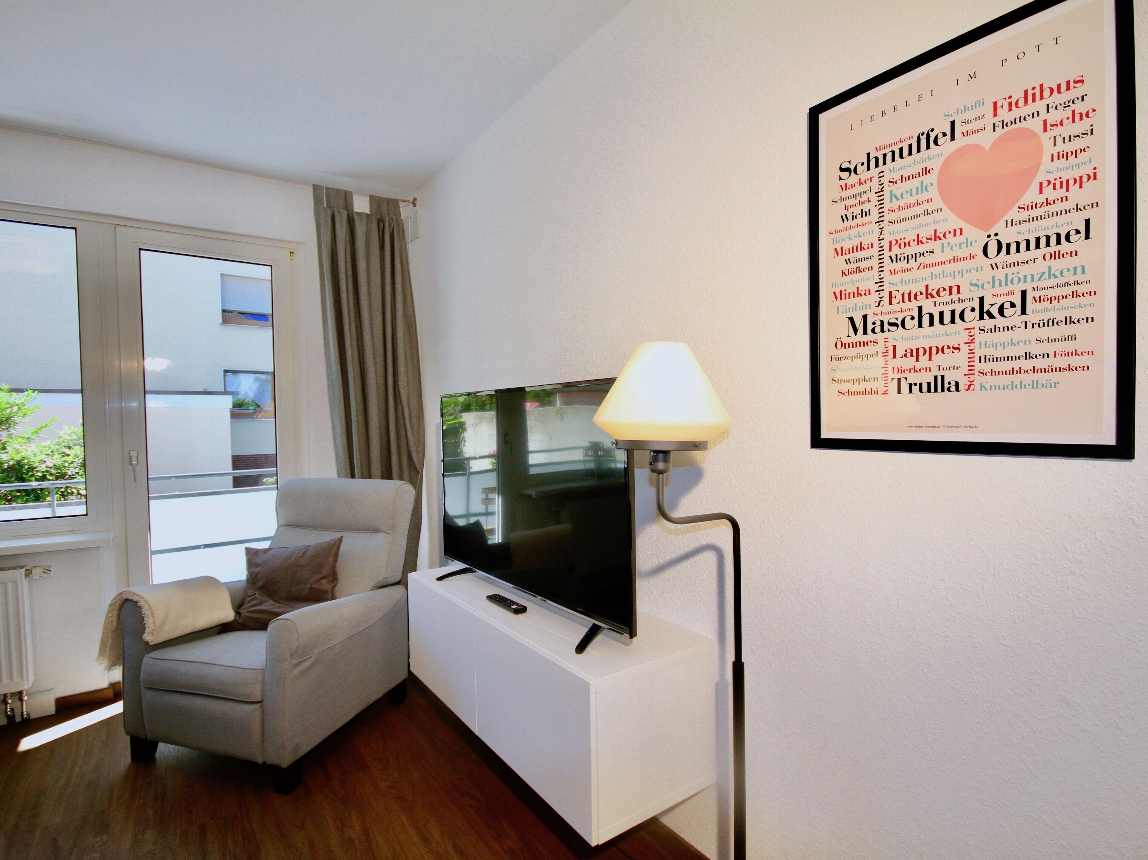 A modern living room with a flat-screen TV on a white stand, a grey armchair with a throw blanket, and a framed word cloud on the wall.