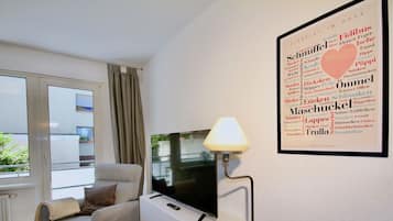 A modern living room with a flat-screen TV on a white stand, a grey armchair with a throw blanket, and a framed word cloud on the wall.