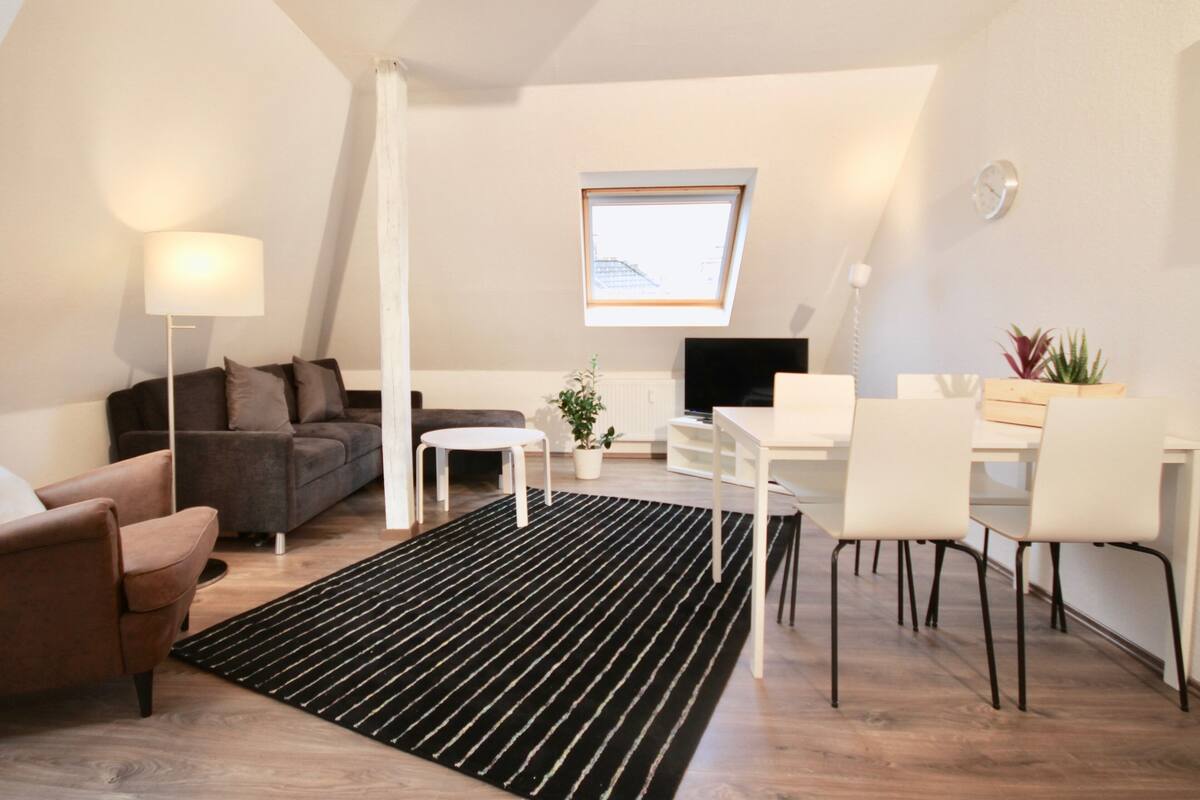 A modern loft apartment with a black and white striped rug, a grey sofa, a small white table, and a white dining area with chairs.