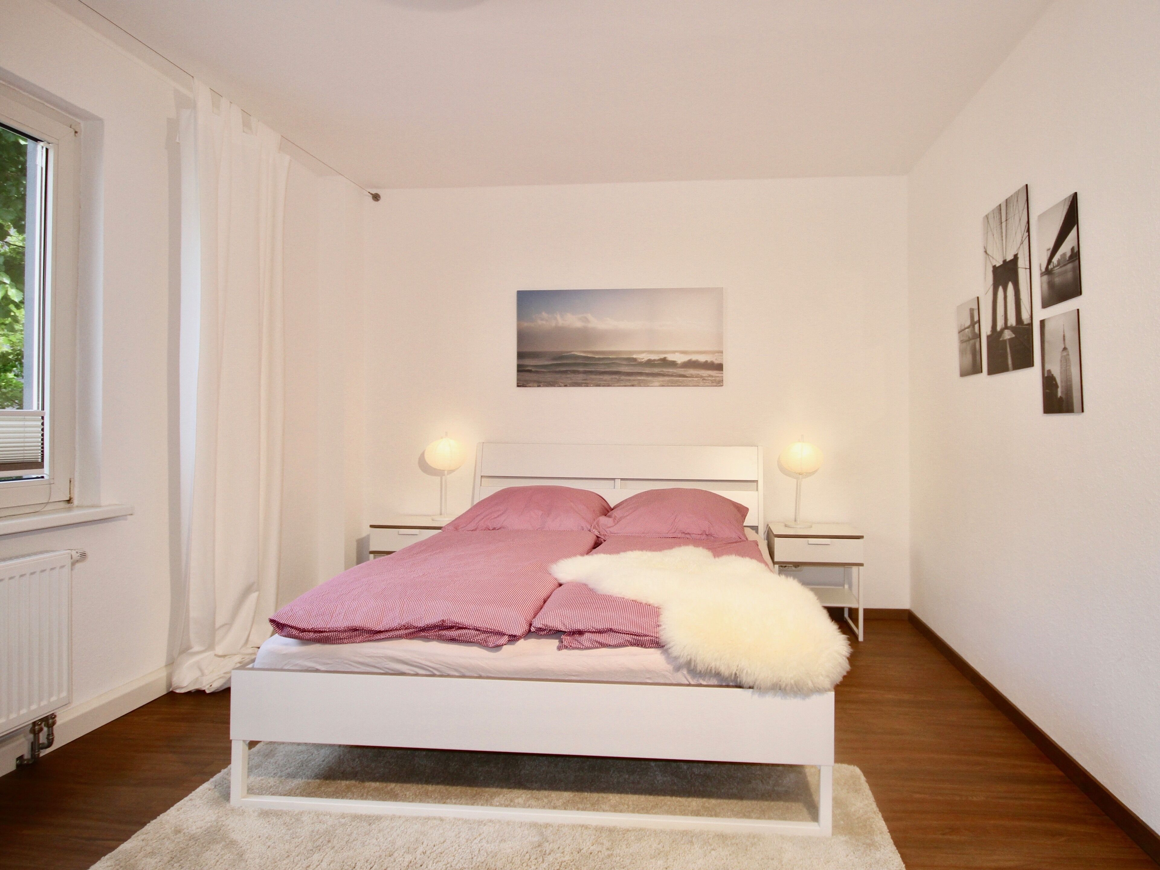 A bedroom with a bed, two bedside lamps, a window with curtains, a radiator, and a wall with framed pictures.