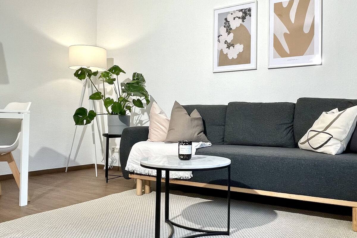 A modern living room with a grey sofa, a small round table, a floor lamp, and a potted plant.