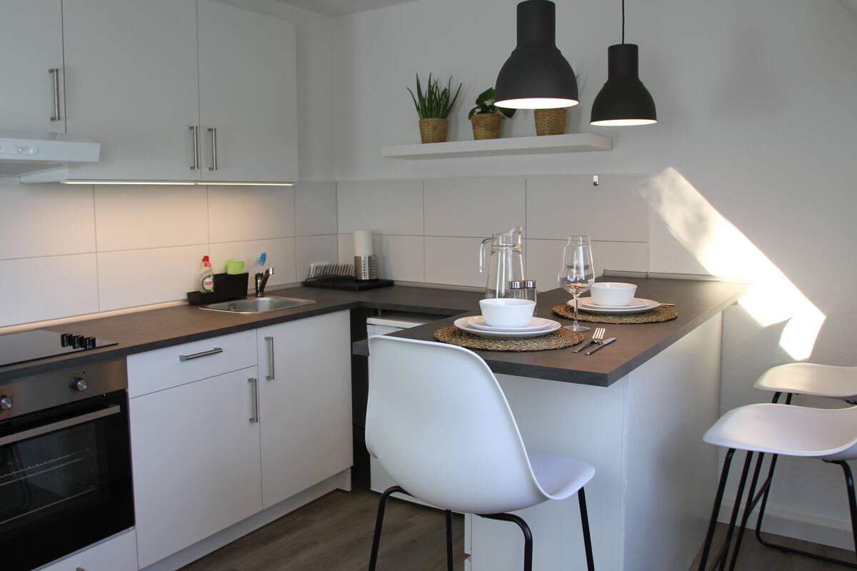 A modern kitchen with white cabinets, a built-in oven, a sink, and a dining area with two white chairs and a table set for two.
