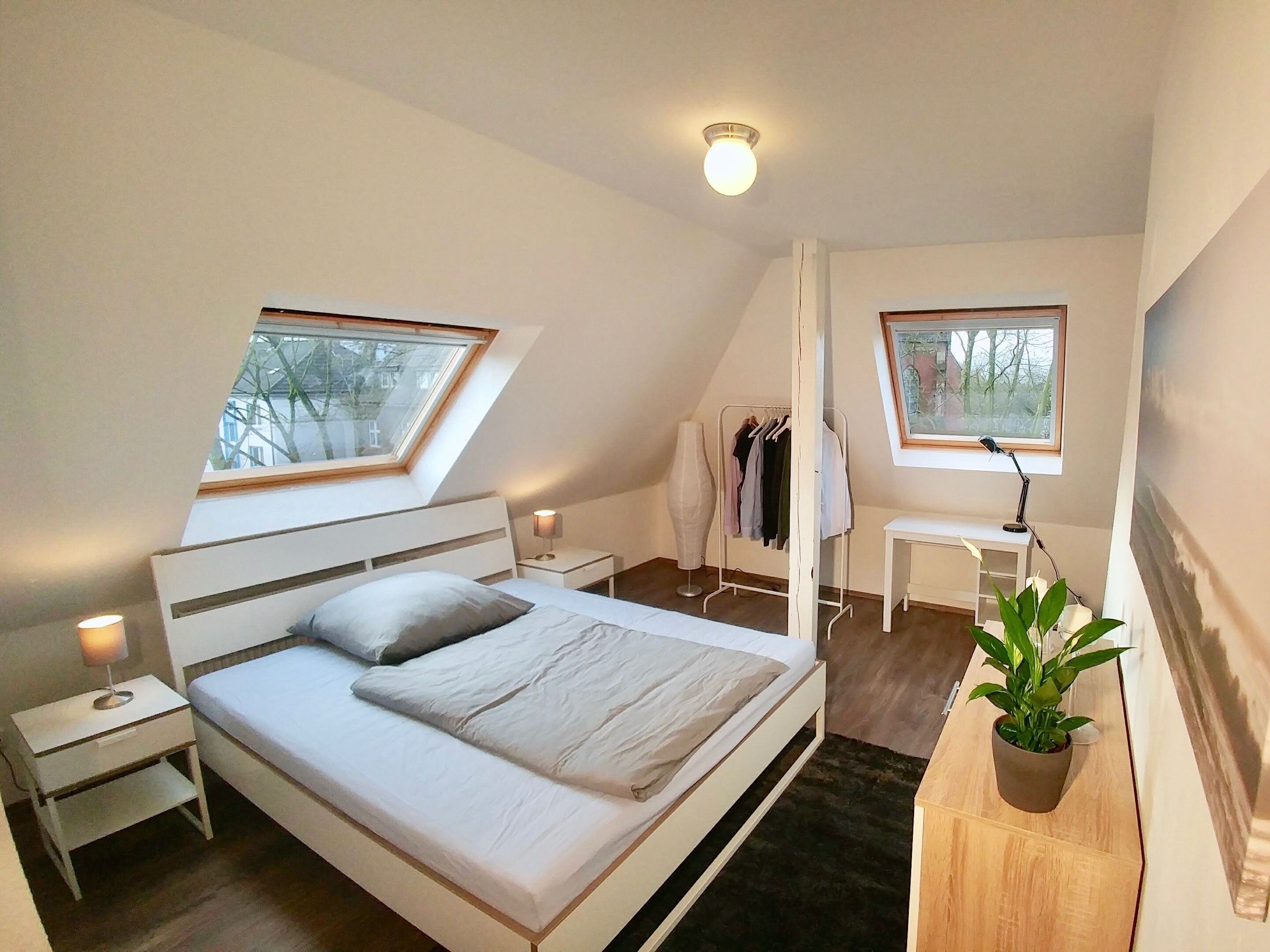 Ein Schlafzimmer mit Bett, Nachttischen, Schreibtisch, Stuhl, einer Zimmerpflanze und einem Fenster.