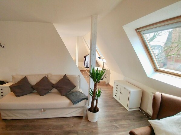 A modern, compact living space with a sloped ceiling, a sofa with cushions, a potted plant, a radiator, and a window with a view of buildings. - Apartmenthaus in der Arnoldstraße (Bochum)