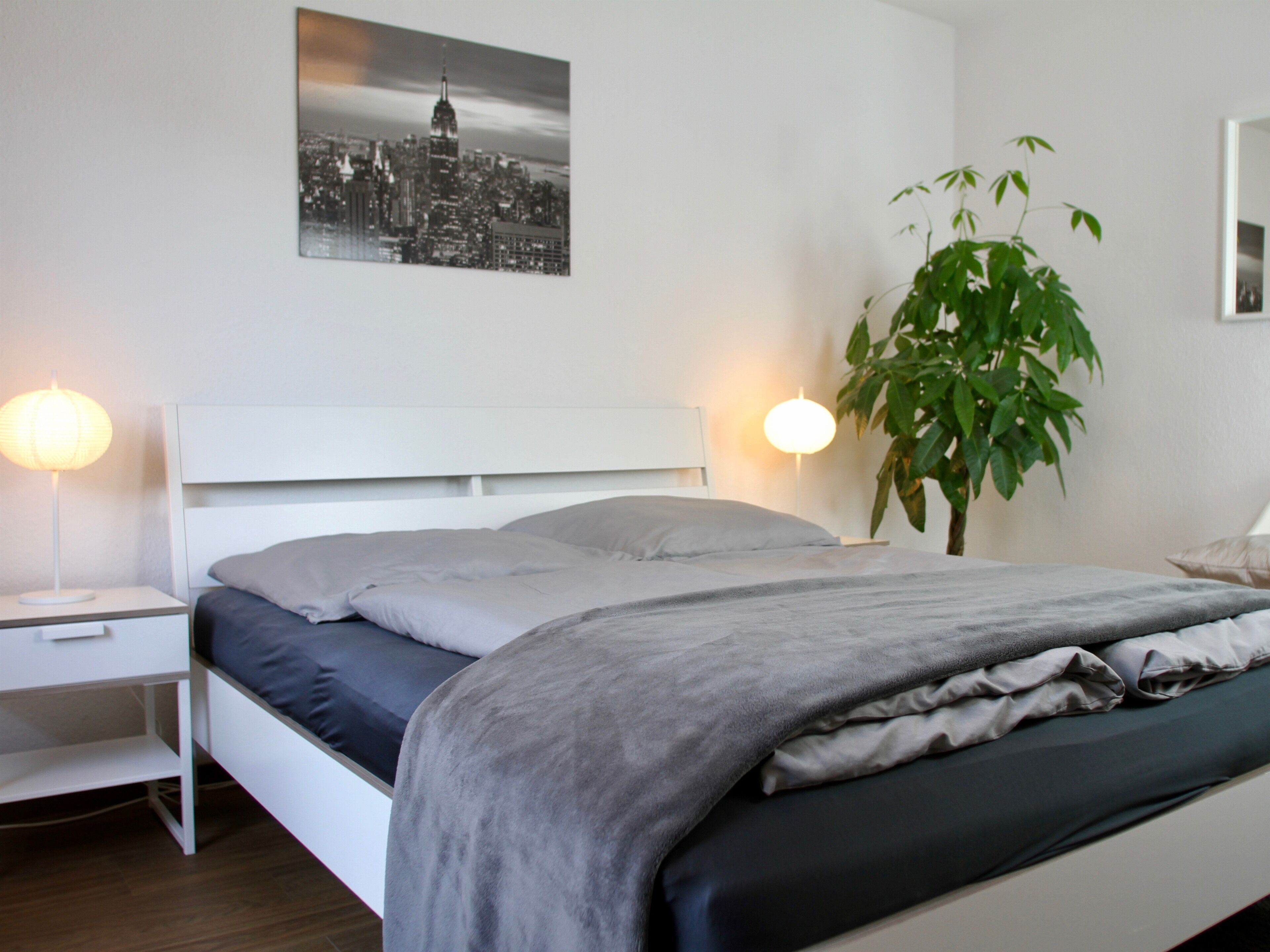 A modern bedroom with a bed, bedside tables, a lamp, and a cityscape painting on the wall.