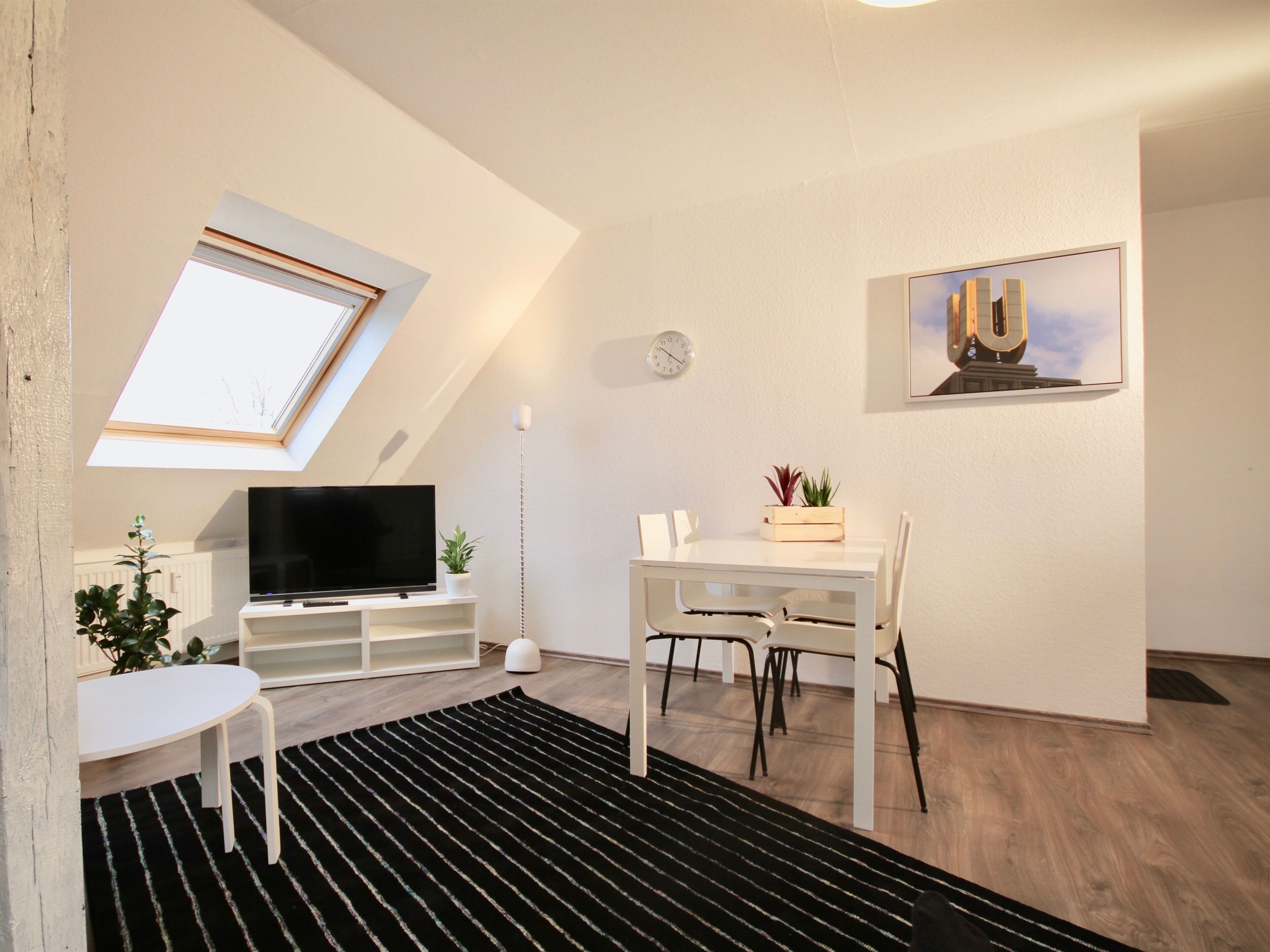 Una sala de estar moderna con un televisor de pantalla plana, una mesa de comedor y sillas. Hay un tragaluz, un reloj en la pared y un cuadro enmarcado sobre la mesa.