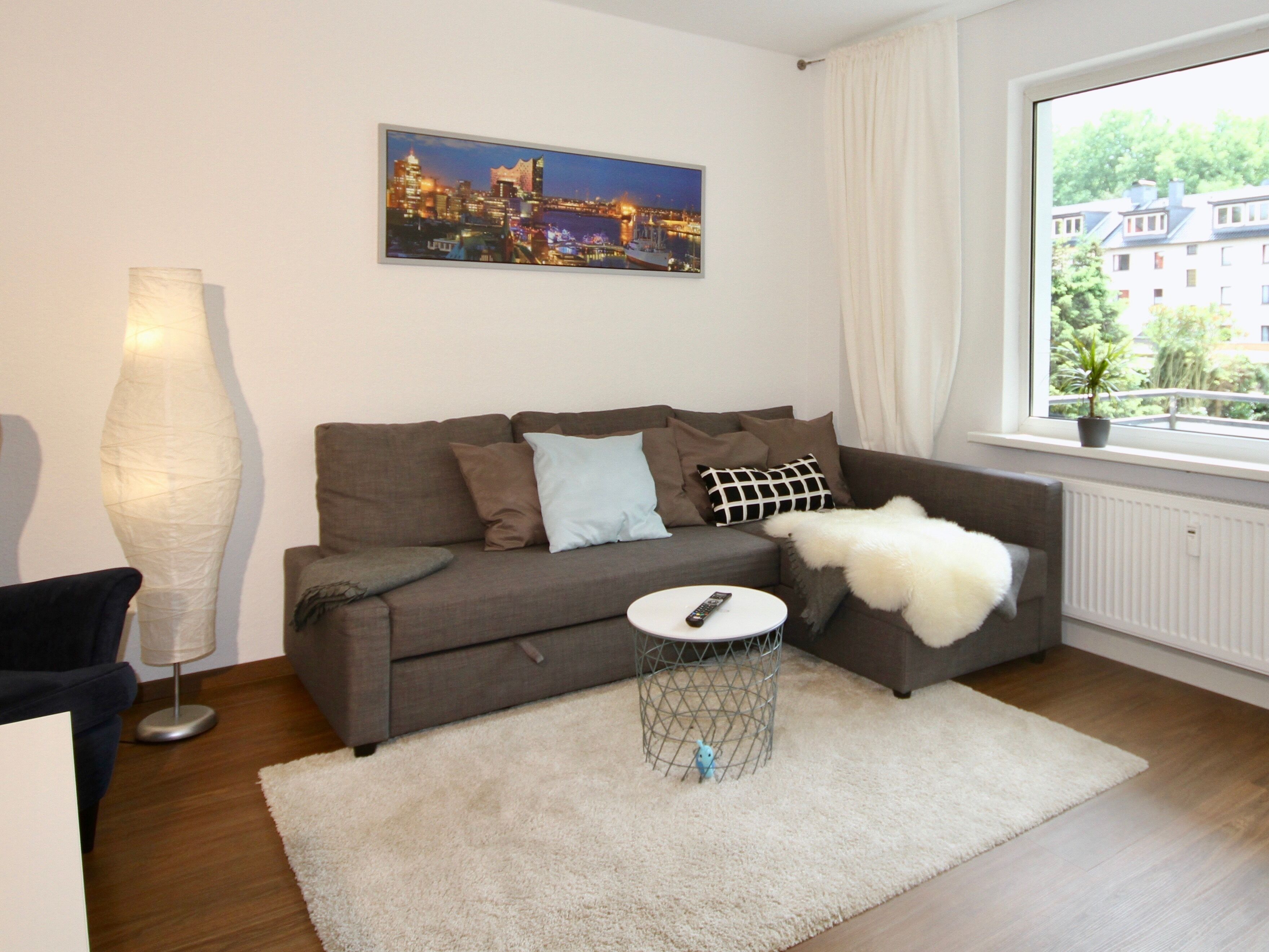 Een moderne woonkamer met een grijze bank, een witte plaid en een stadslandschap schilderij aan de muur.