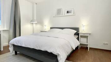 Een net opgemaakt bed met witte beddengoed, twee nachtkastjes met lampen, een ingelijst schilderij aan de muur en een raam met gordijnen.