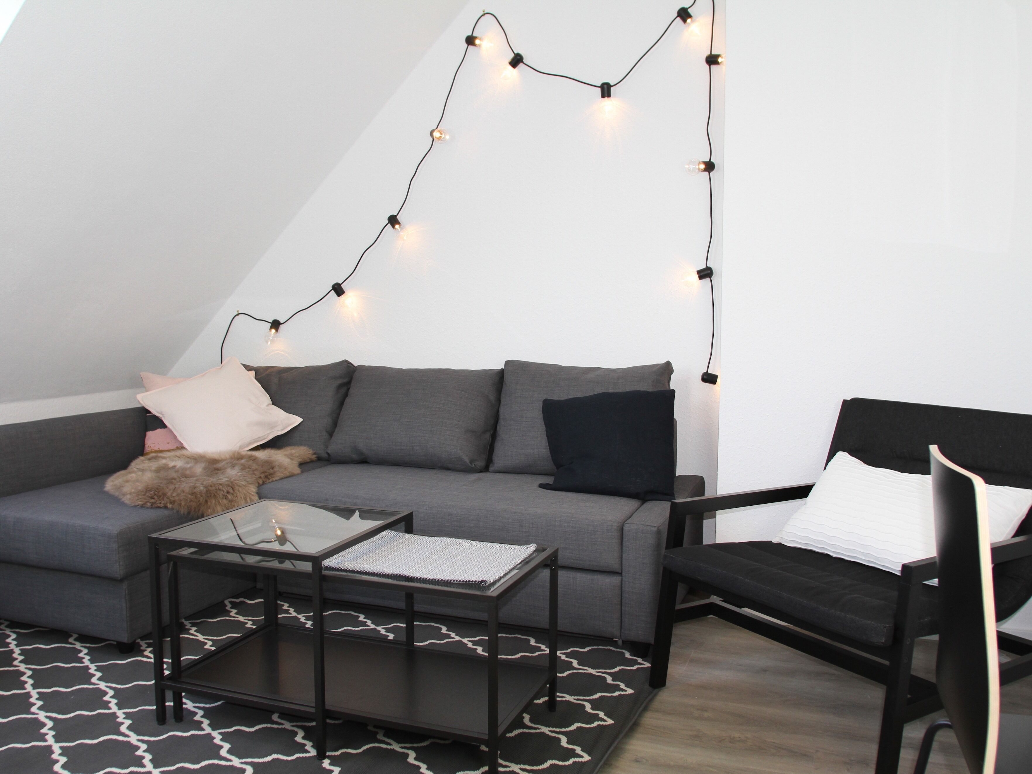 Camera da loft moderna con un divano grigio, una sedia nera, un tavolino in vetro e luci a stringa sul soffitto.